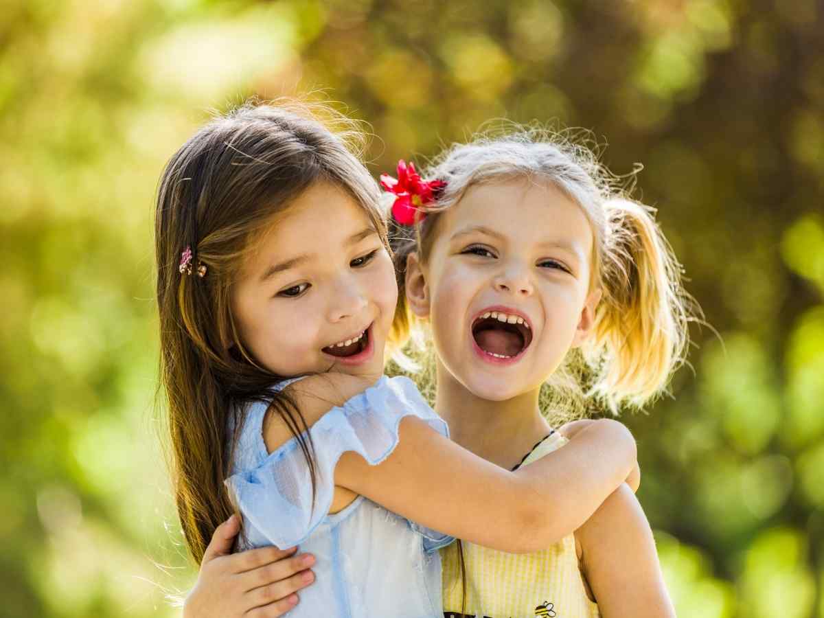 Foto de duas crianças meninas sorridentes se abraçando. Elas estão em área com árvores no fundo.
