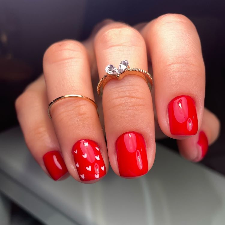 Mãos com unhas curtas esmaltadas em vermelho brilhante, uma delas decorada com pequenos corações brancos. Dedos adornados com anéis delicados, incluindo um com detalhe de coração em cristal. Fundo desfocado em tons neutros, destacando o design romântico.