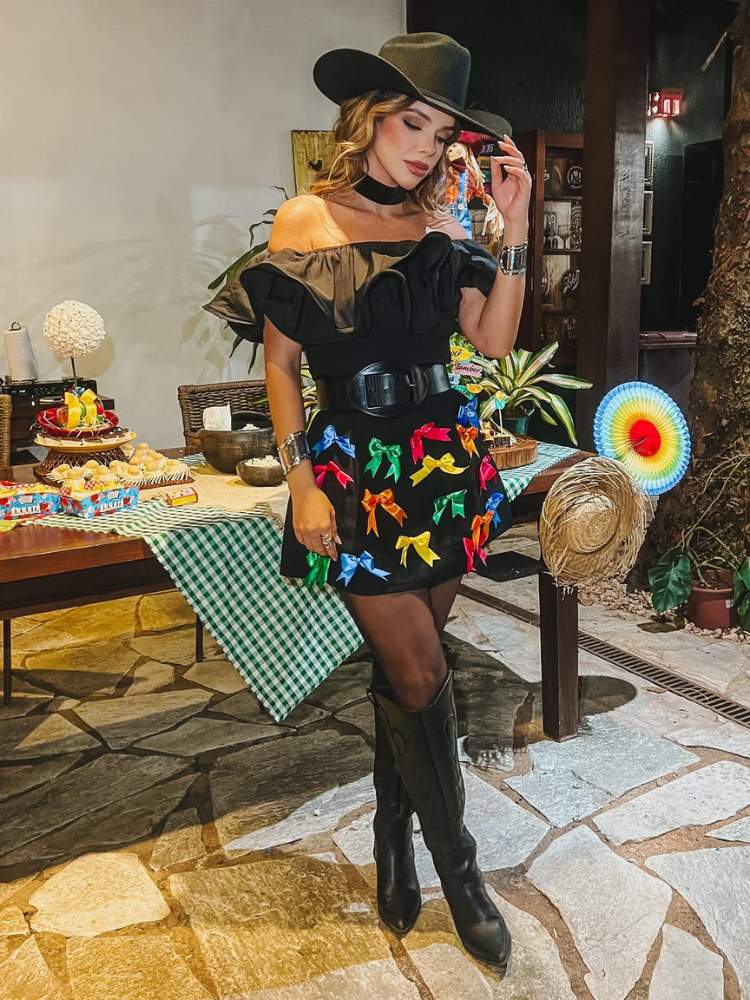 Mulher de pele clara com look preto, saia com fitas coloridas, bota e chapéu country 