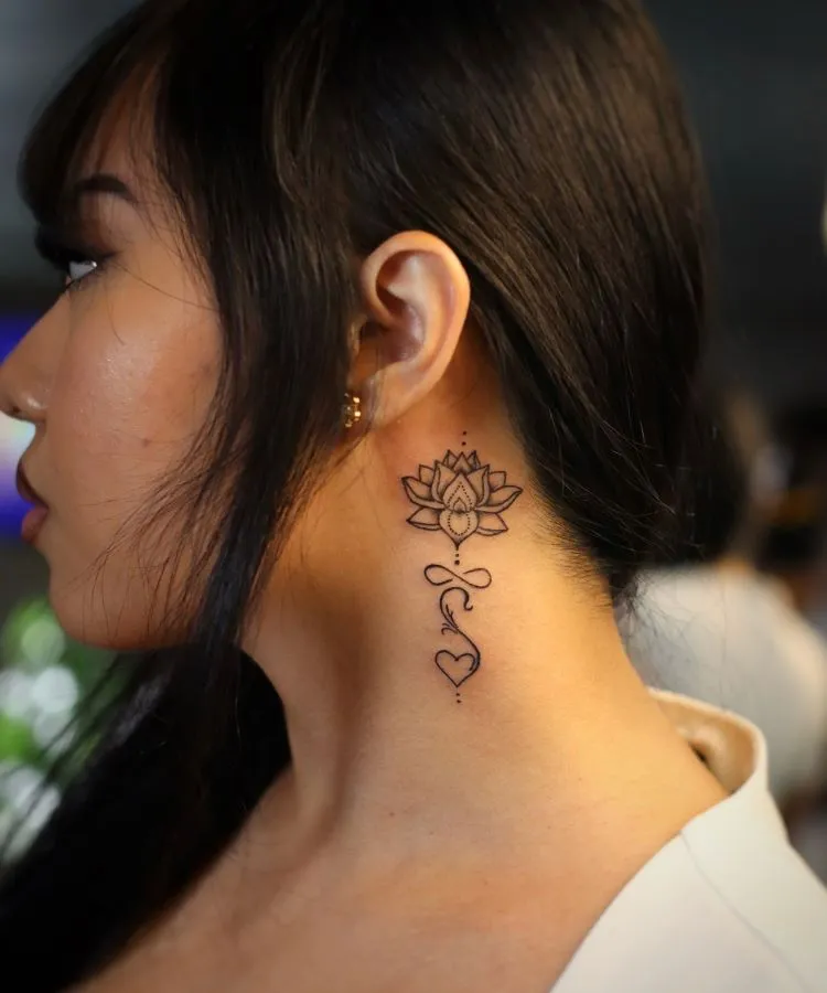 A imagem mostra uma pessoa de perfil com cabelo escuro e liso. Ela tem uma tatuagem no pescoço de uma flor de lótus detalhada, com uma linha que desce até um coração. A tatuagem é delicada e em preto, localizada atrás da orelha e estendendo-se pela lateral do pescoço.




