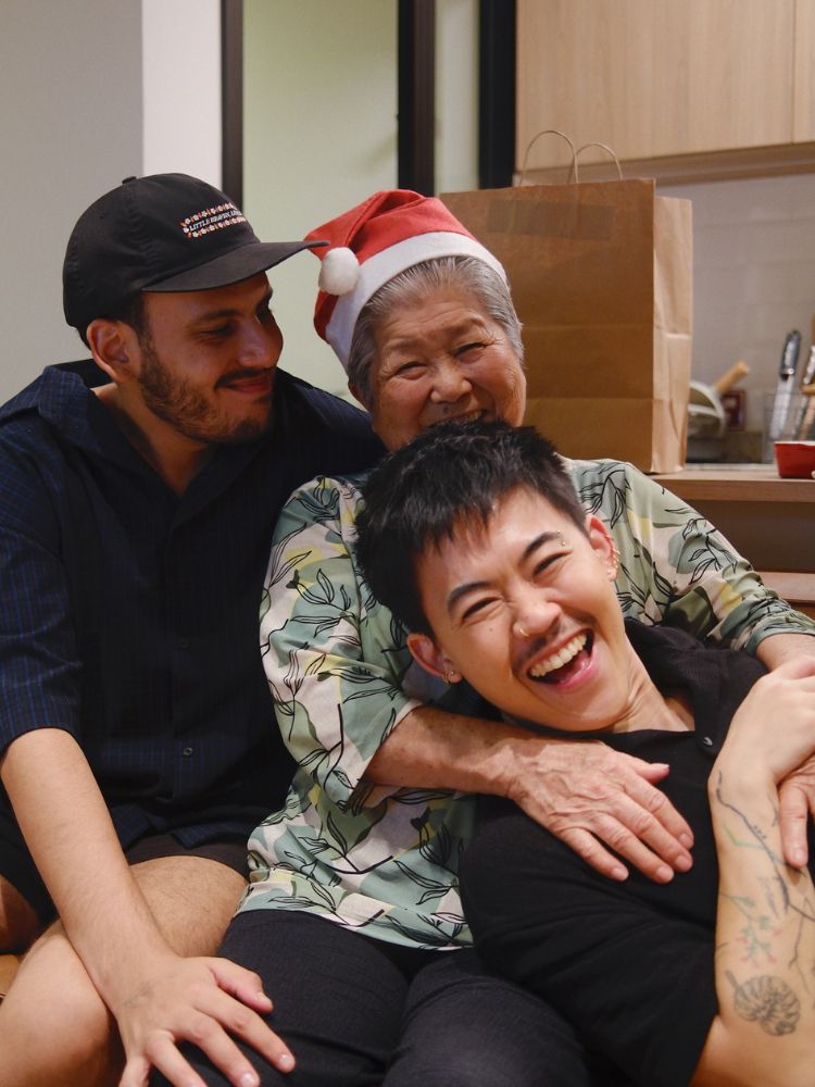Léo de camiseta preta no colo se sua avó de roupa estampada verde com gorro do papai Noel, sendo abraçada por Alan, também de roupa preta 