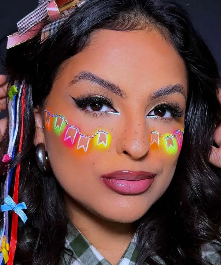 Uma mulher com maquiagem vibrante de Festa Junina, apresentando delineado gráfico preto e branco, lábios rosados e um colar de bandeirinhas neon desenhado no rosto. Seu cabelo escuro é adornado com fitas coloridas, e há um chapéu de palha com laço xadrez.