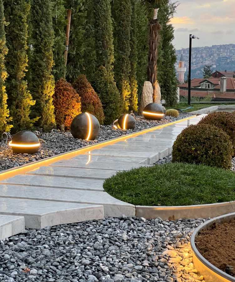 Caminho de mármore iluminado por lâmpadas esféricas modernas com detalhes dourados, cercado por brita cinza e jardins com grama e arbustos podados. Árvores altas proporcionam privacidade, enquanto ao fundo se vê um cenário urbano ao entardecer.