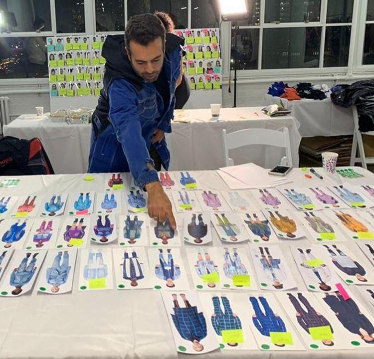 João Foltran atrás de uma grande mesa cheia de desenhos de combinações de roupas. Ele aponta para um desenho no centro da mesa.