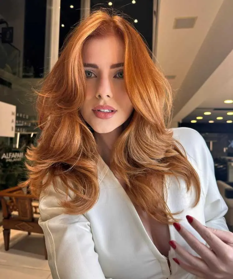 Mulher com cabelos longos, ruivos e volumosos, estilizados em camadas suaves. Ela veste uma blusa branca elegante, com maquiagem leve e batom rosado. Ambiente iluminado com decoração moderna e detalhes em tons neutros ao fundo, criando um contraste sutil.