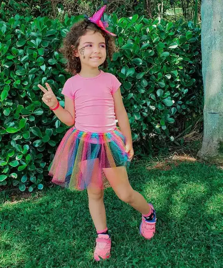 Garota com fantasia de bruxa infantil. Cores predominantes: rosa, azul e amarelo. Usa: camiseta, saia de tule colorida, tênis, tiara de mini chapéu