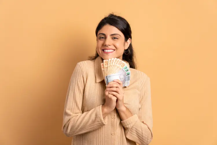 A imagem mostra uma mulher sorridente, de cabelos escuros e longos, segurando notas de euro em leque. Ela veste uma blusa bege, com um fundo laranja claro ao redor.