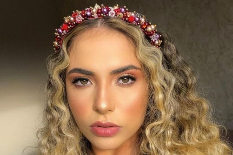 Mulher de pele clara com cabelo loiro escuro ondulado e com tiara de pérolas e miçangas vermelhas e cintilantes