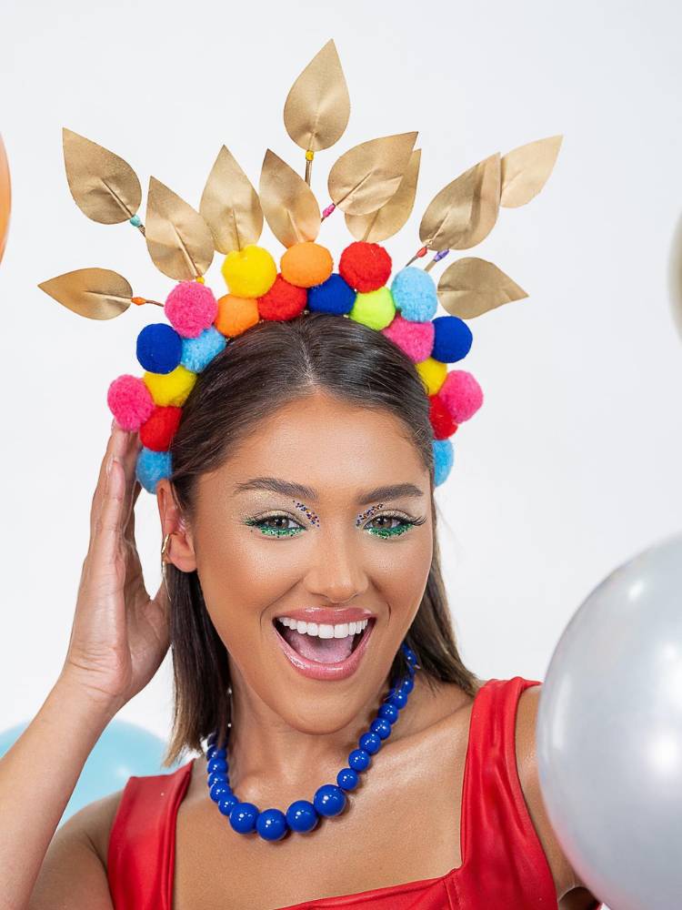 Mulher de pele morena e cabelo escuro maquiada, usando roupa vermelha, colar azul e tiara com pompons coloridos e folhas douradas.