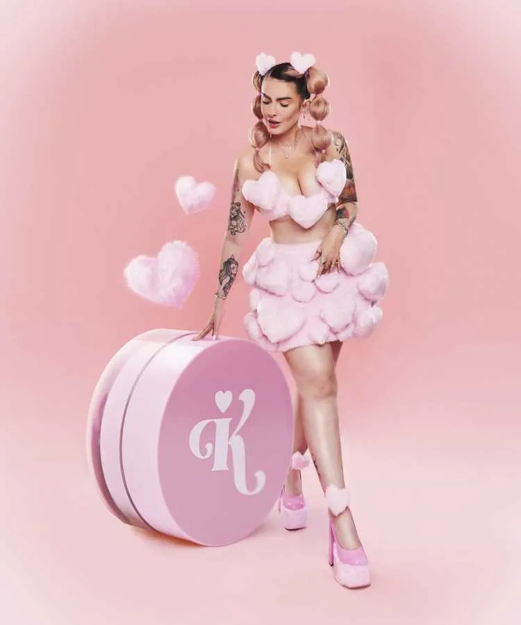 Karen Bachini vestida em roupas temáticas com corações de pelúcia rosa posa em fundo totalmente rosa. Ela usa sapatos combinando, penteado com corações e acessórios pink. Ao lado, um grande cilindro rosa com letra "K" branca destacada.