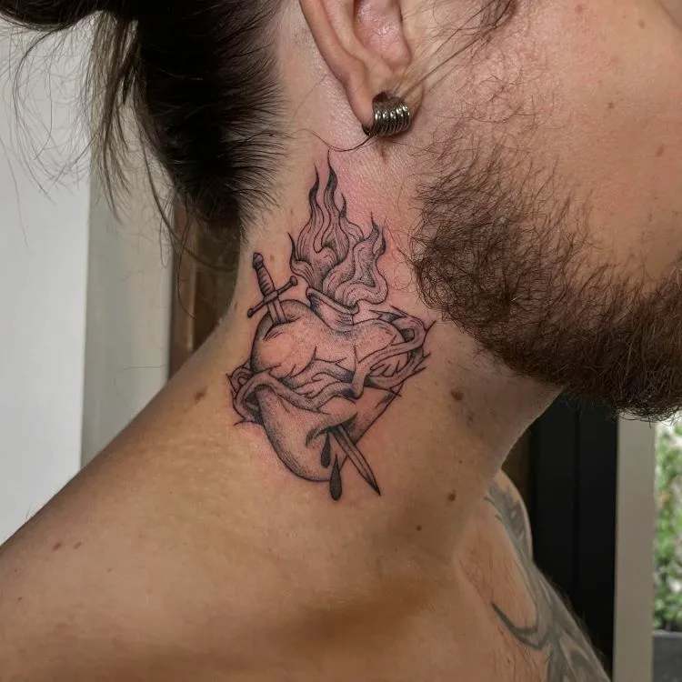 A imagem mostra uma pessoa de perfil com cabelo preso e barba. Ela tem uma tatuagem no pescoço de um coração anatômico com chamas saindo do topo e uma espada atravessando-o. A tatuagem é detalhada em preto e cobre a lateral do pescoço até a parte de trás da orelha.


