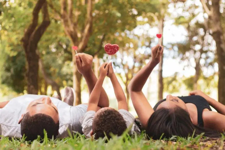 Família deitada em um gramado sob árvores ao ar livre. Pai, filho e mãe seguram pirulitos de coração no alto, simbolizando amor e união. A luz natural e o cenário verde proporcionam um ambiente leve, carinhoso e descontraído.