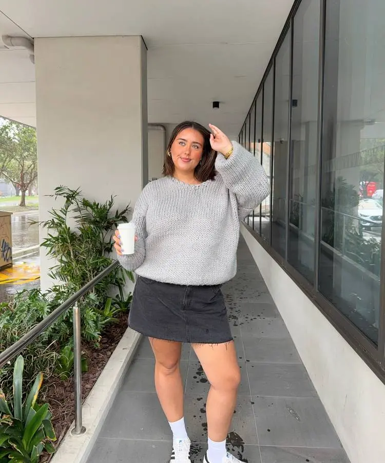 Uma mulher sorridente com cabelo castanho e ombros largos veste um suéter cinza de tricô oversized e uma minissaia jeans preta. Ela segura um copo branco e usa meias brancas com tênis brancos, caminhando em uma passarela externa com vegetação ao fundo.