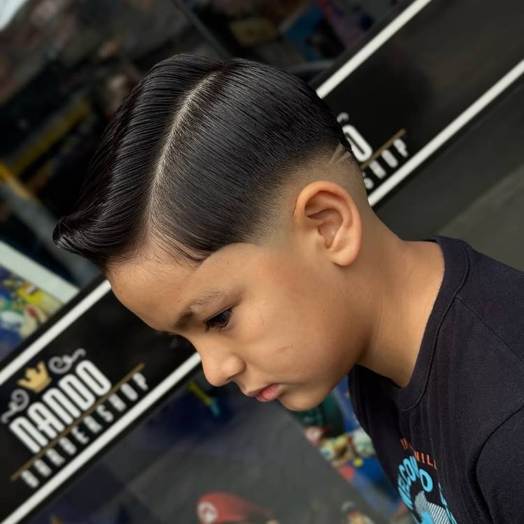 Um menino com um **corte de cabelo na lateral** e um degradê (fade) nas laterais e na nuca. O cabelo do topo é escuro e liso, penteado com uma linha lateral bem marcada e um design de linhas raspadas na nuca.