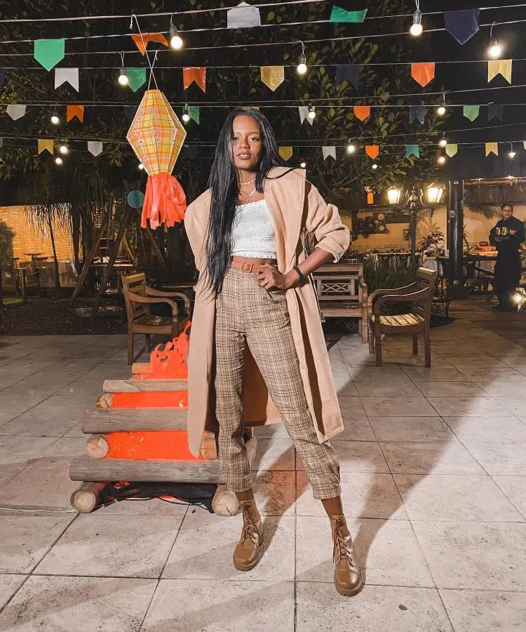 Mulher negra confiante, cabelo liso longo, casaco bege aberto sobre camiseta branca brilhante e calças xadrez, mão no quadril. Ambiente noturno com luzes de corda, bandeiras coloridas e fogueira artificial em evento festivo ao ar livre.





