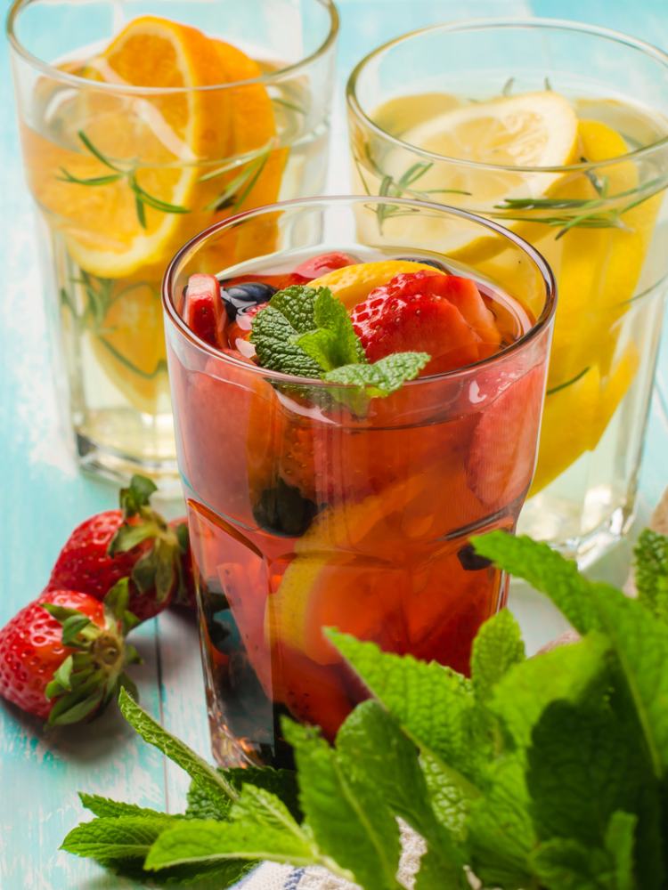 3 copos com água saborizada, frutas como morango, laranja e limão, além de hortelã