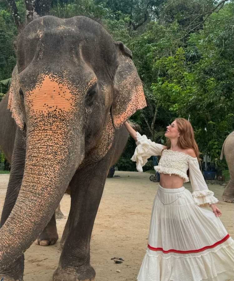 Mari Maria, influenciadora digital, posa ao lado de um elefante em ambiente natural. Ela veste um conjunto branco com detalhes delicados, enquanto olha para o animal, transmitindo uma conexão pacífica e harmoniosa com a natureza ao fundo.
