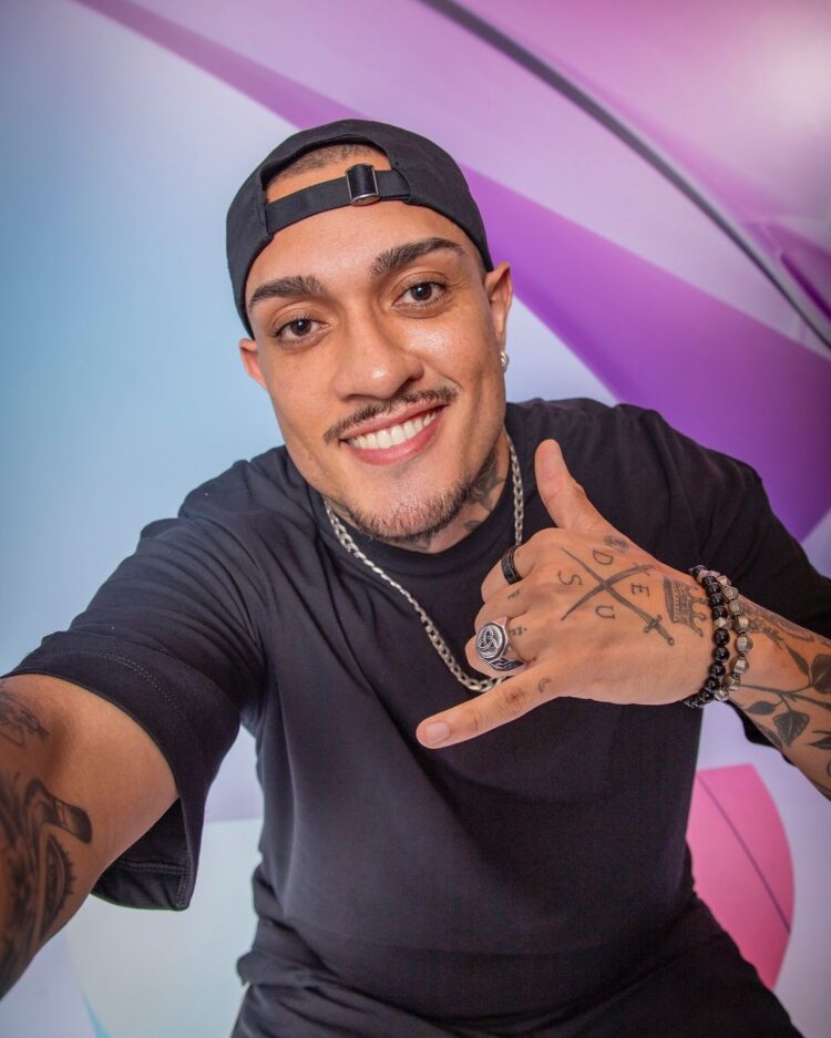 Mc Bin Laden de blusa preta, boné, corrente prateada e fazendo seu símbolo de "tá tranquilo, tá favorável"
