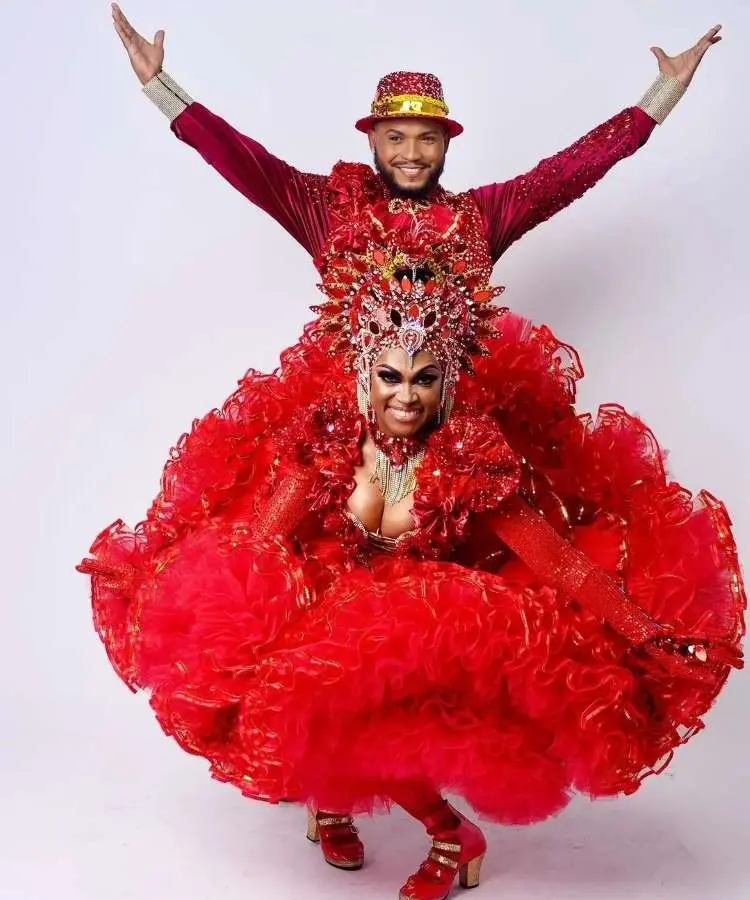 Imagem de um casal de dançarinos da quadrilha em festa junina, ambos com trajes vermelhos e brilhantes. A mulher, em destaque, usa um vestido volumoso com coroa e o homem, atrás, tem os braços abertos, celebrando a alegria e o folclore. Fontes