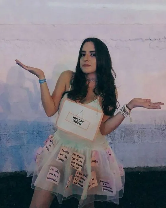 Foto de mulher de pele clara com look branco, plaquinha "papel de trouxa" e post-its na saia com frases 
