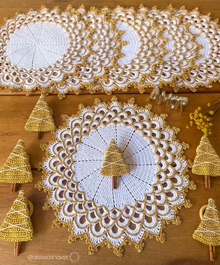 Sousplats de crochê branco com bordas douradas dispostos sobre mesa de madeira. Pequenos enfeites de árvore de Natal dourados complementam a decoração sofisticada e festiva, refletindo um tema natalino elegante.