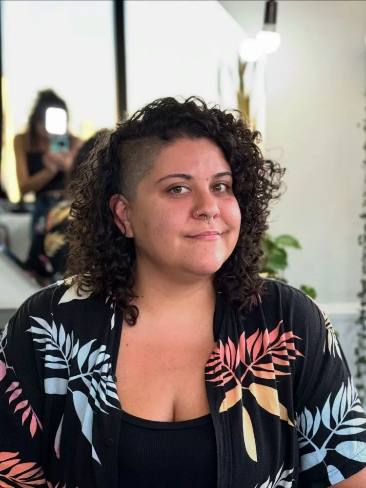 Mulher de pele clara e rosto redondo usando camisa de fundo preto e flores coloridas e cabelo cacheado sidecut