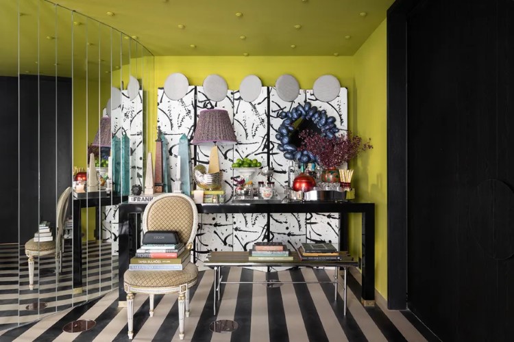 A imagem mostra um interior de sala com estilo moderno e artístico. Destaca-se uma parede amarela com grafite de rostos em preto e branco, uma parede preta lisa, e o piso é xadrez em preto e branco. O mobiliário inclui uma mesa preta com objetos decorativos, cadeiras estampadas, e luminárias pendentes.

