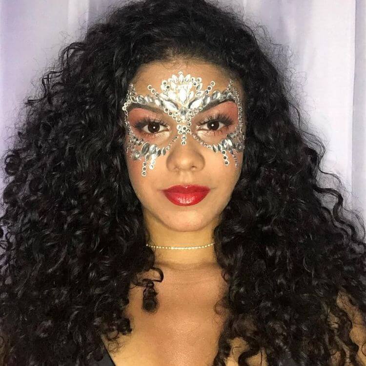 Mulher de pele negra usando maquiagem terrosa, batom vermelho e pedrarias prateadas formando máscara 