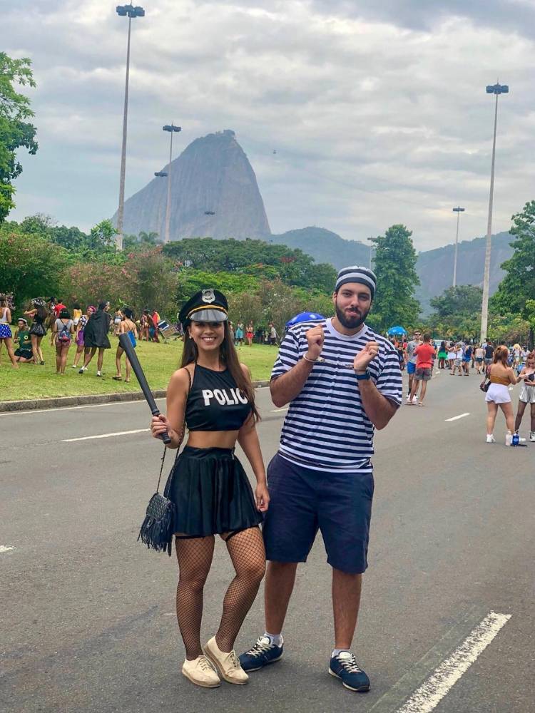 Mulher de pele clara suando cropped preto escrito "policial", saia preta e chapéu de policial, além de cacetete. Ao lado, homem de camiseta listada branca e preta e bermuda