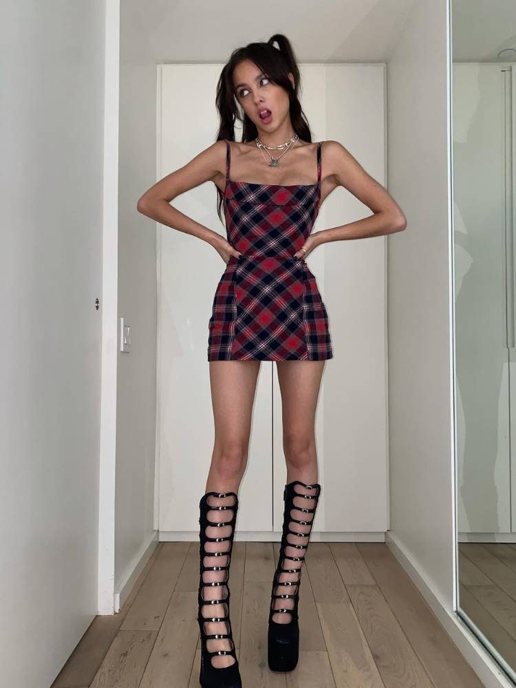 Olivia Rodrigo usando vestido curto xadrez, bota preta de cano longo e penteado com amarrações aleatórias