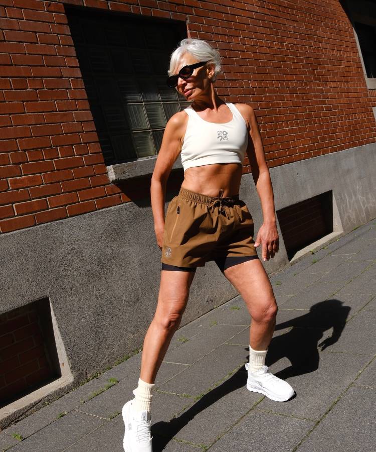 Uma mulher atlética de cabelo grisalho e óculos escuros veste um top branco, shorts marrons com calção preto por baixo, meias brancas caneladas e tênis brancos. Ela posa em uma rua pavimentada, com um prédio de tijolos à sua direita.
