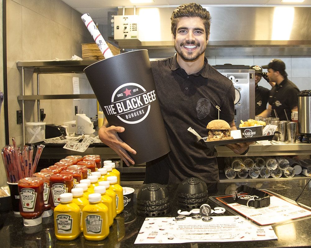 Caio Castro na sua hamburgueria, de uniforme preto e segurando combo