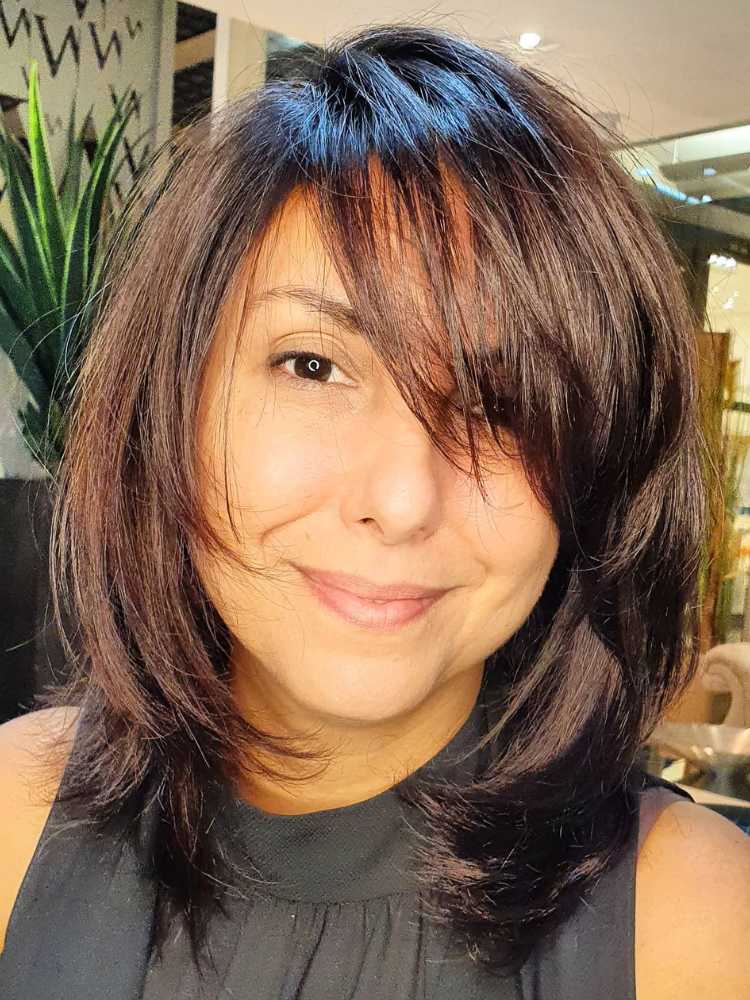 Mulher de pele morena usando regata preta, sorrindo, com corte de cabelo chanel com franja lateral repicada