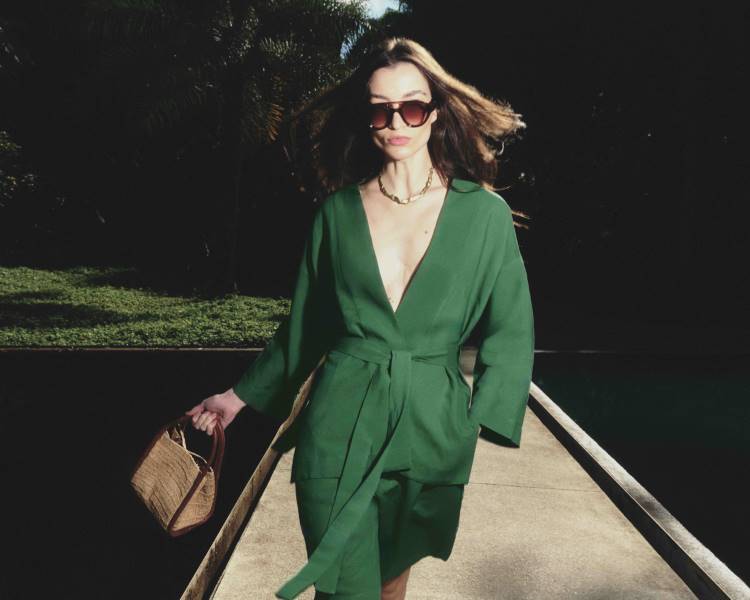 Mulher de óculos de sol e cabelos ao vento, caminha em passarela estreita sobre a água. Veste um conjunto de blazer e shorts verdes, amarrado na cintura, e carrega uma bolsa de palha.Coleção Lenny Niemeyer + Inhotim.