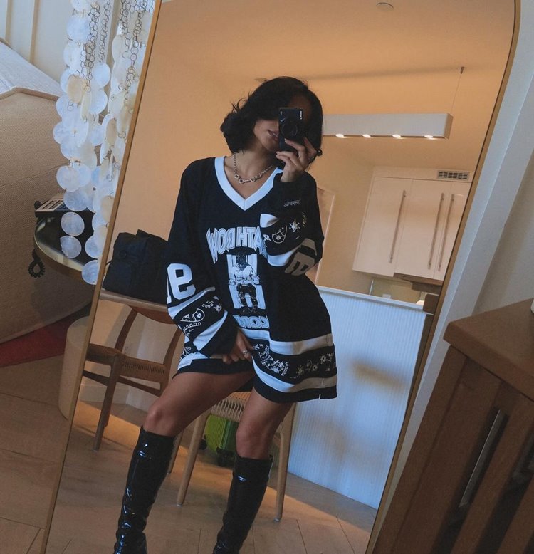 Becky G com corte bob está em frente a um espelho, vestindo uma camiseta oversized preta com estampas brancas de estilo esportivo. Ela usa botas de cano alto pretas e segura o celular para tirar a foto. O ambiente é uma sala bem iluminada. 