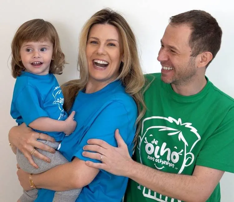 Uma mulher sorridente em uma camiseta azul segura uma criança pequena com cabelos castanhos e camiseta azul. Um homem em uma camiseta verde "de olho nos olhinhos" sorri ao lado. Nomes diferentes de filhos de famosos: esta família demonstra afeto e união.

