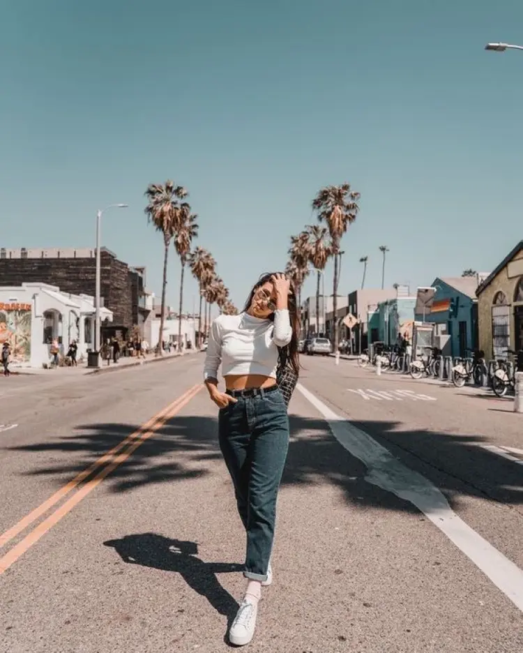Frase para foto pensativa mulher branca no meio da rua usa óculos escuros, veste calça jeans de cintura alta com cinto preto e blusa cropped branca com manga e gola altao Frase para foto pensativa mulher branca