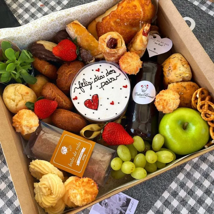 A imagem mostra uma cesta de Dia dos Pais repleta de itens comestíveis variados, como frutas frescas, pães, doces e uma garrafa de bebida. Há um bolo com a frase “Feliz dia dos Pais”. A diversidade e apresentação tornam a cesta visualmente atraente.


