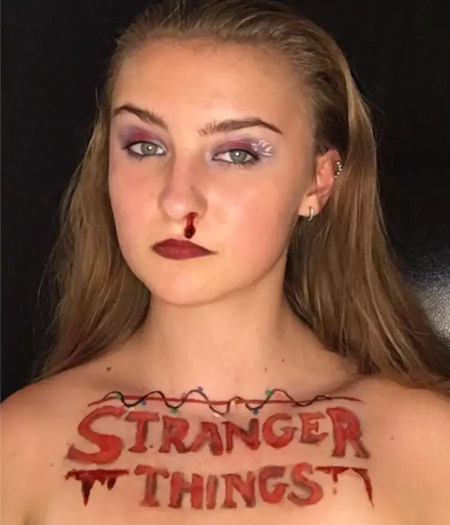 Mulher de pele clara com sombra vermelha e branca, nariz com sangue falso e colo escrito "stranger things"