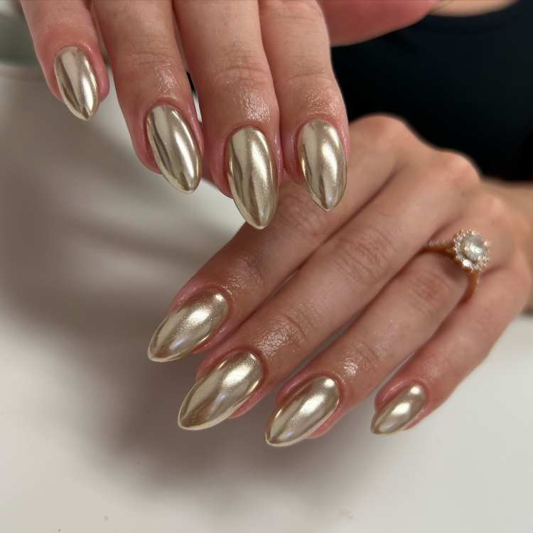 Mãos femininas com unhas no formato amendoado, pintadas em tom dourado metálico brilhante. O acabamento reluzente reflete luz suavemente, destacando elegância. Uma das mãos usa um anel com pedra central, compondo o visual sofisticado e alinhado às tendências de 2025.