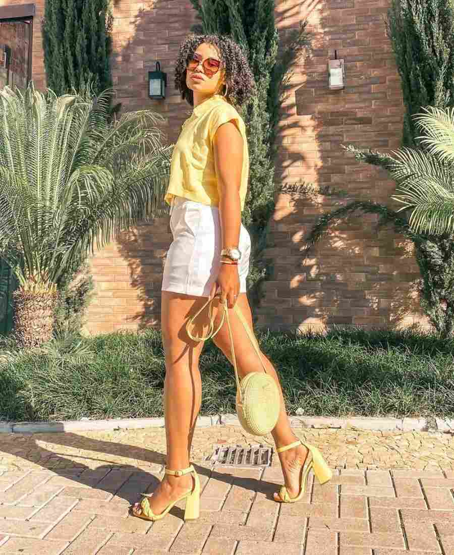 Imagem com fundo de parede com plantas. No centro, mulher usando roupas amarela: camiseta amarela + short branco + bolsa amarela + sandália de salto amarela.