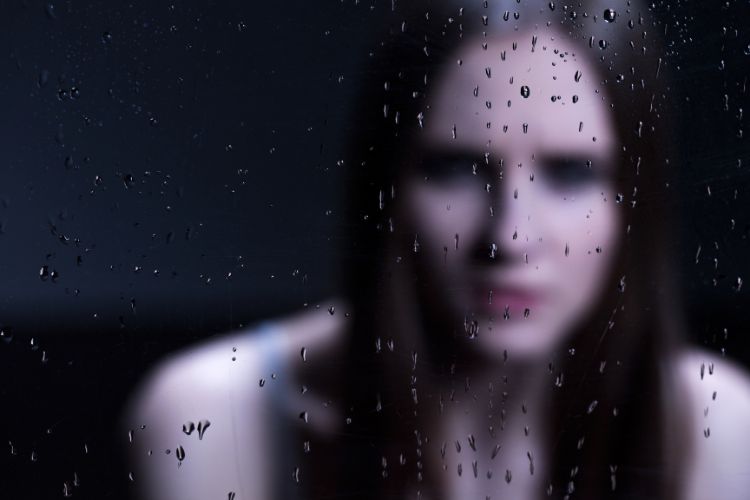 Uma mulher desfocada aparece atrás de um vidro com gotas de chuva, transmitindo melancolia. A imagem simboliza emoções complexas como o ciúme, frequentemente analisadas na psicanálise, que explora inseguranças e sentimentos refletidos em situações de vulnerabilidade emocional