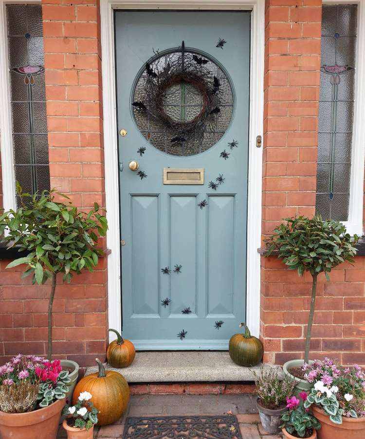 Entrada de casa com parede de tijolos à vista, porta cinza com guirlanda seca e brinquedos de aranha colados, vasos de flores e abóboras