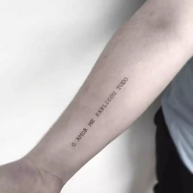 A imagem mostra uma tatuagem minimalista no antebraço com a frase "O amor me explicou tudo", escrita em letras maiúsculas e simples. A pele é clara, e o fundo neutro destaca o design discreto e significativo da tatuagem.