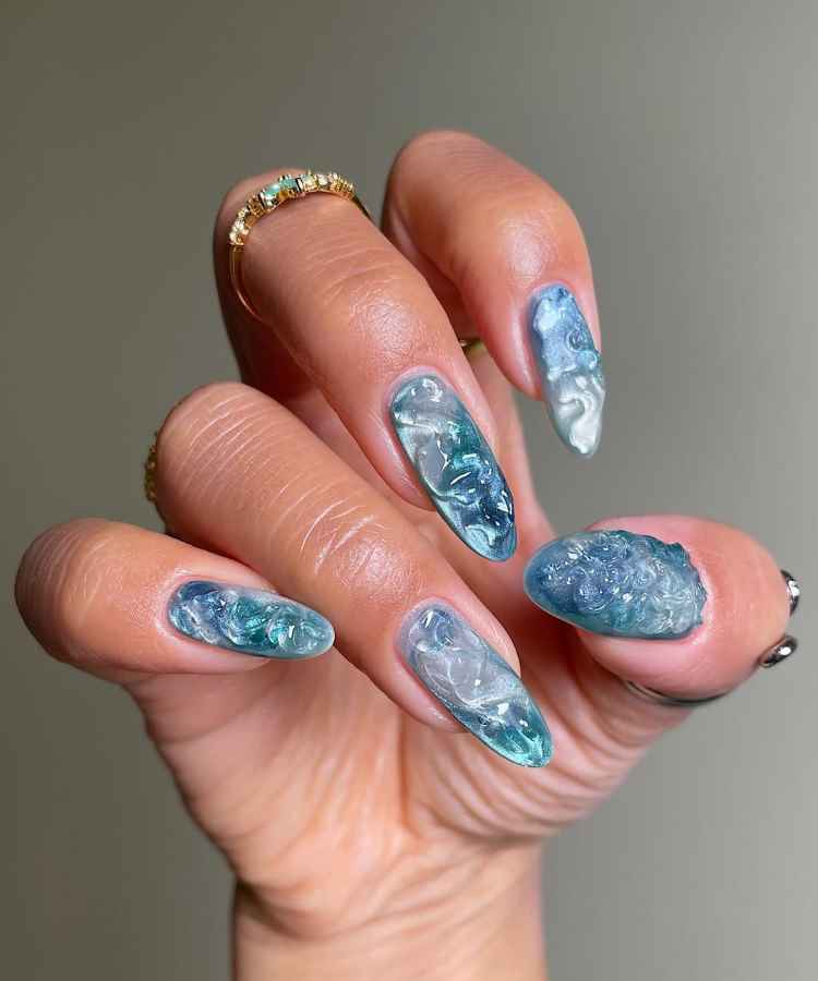 Unhas compridas com design em azul e branco, criando um efeito marmorizado em textura 3D. A mão usa anéis dourados delicados, destacando a arte nas unhas. O fundo é cinza, realçando os tons vibrantes e o estilo sofisticado das unhas.