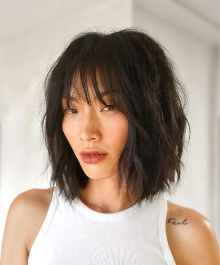 Mulher com cabelo médio escuro em corte bob bagunçado, com franja desfiada cobrindo a testa. Ela veste regata branca e tem tatuagem no ombro esquerdo. O fundo é iluminado e desfocado, destacando seu rosto expressivo.