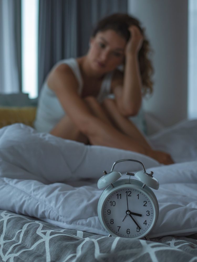 Um despertador analógico branco está em foco no primeiro plano, enquanto uma mulher desfocada senta-se na cama no fundo. Ela parece estar frustrada ou com insônia.