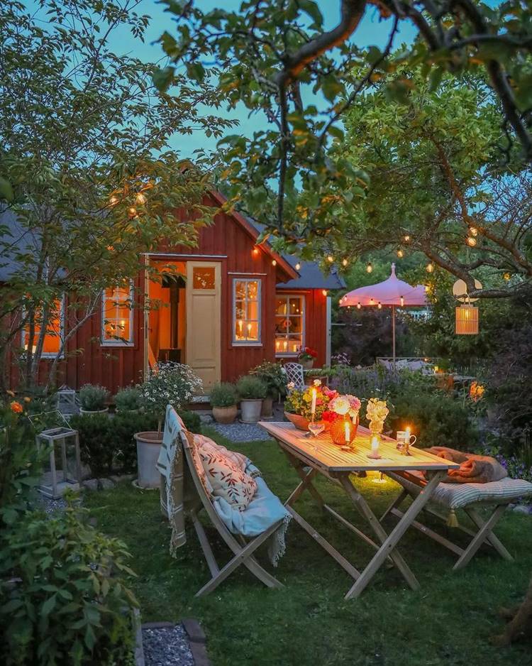 Imagem de fachada de casa de madeira vermelha. Jardim com muitas plantas e árvores possui uma mesa com cadeiras com cobertores e almofadas, decorada com flores em vasos e também velas. Há um varal de lampadas por todo ambiente.
