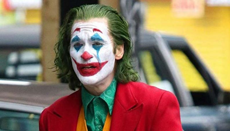Zoom em personagem Coringa, usando cabelo verde, rosto branco, sobrancelha vermelha, nariz vermelho, boca vermelha, sombra azul 
