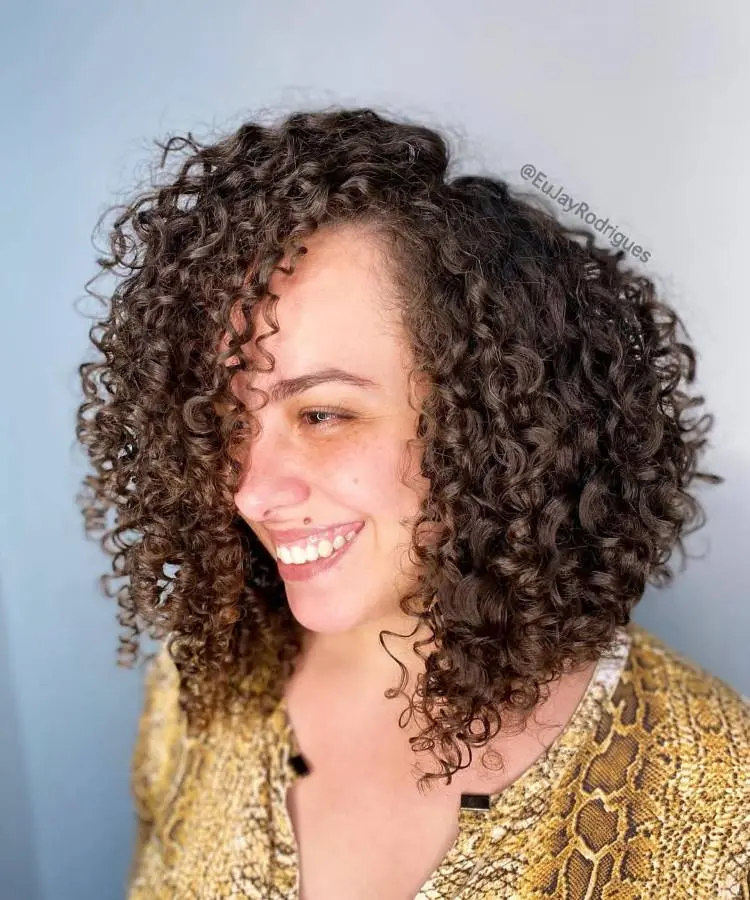 Uma mulher sorridente com cabelos cacheados volumosos, em tons de castanho, olhando para o lado. Seus cachos caem sobre o rosto e ombros. Ela veste uma blusa com estampa de cobra e o fundo é azul.
